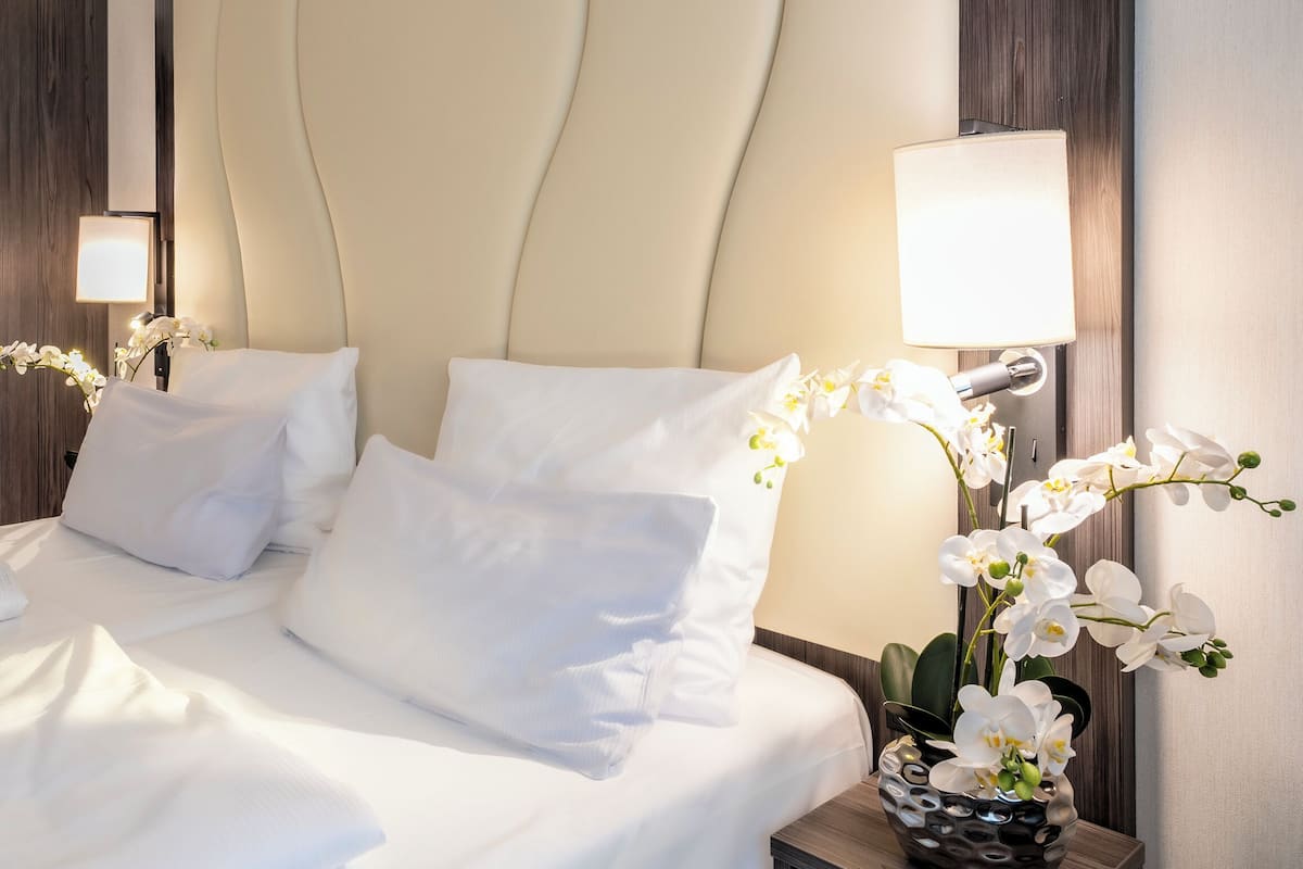 A hotel room with a bed, white pillows, a nightstand with a lamp, and a vase with white flowers.
