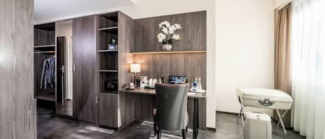 A hotel room with a wooden wardrobe, a desk with a lamp, a chair, a small table, and a suitcase.