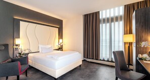 A hotel room with a large bed, two bedside tables with lamps, a chair, and a view of the outdoors through curtains. - PLAZA INN Mannheim (Mannheim)