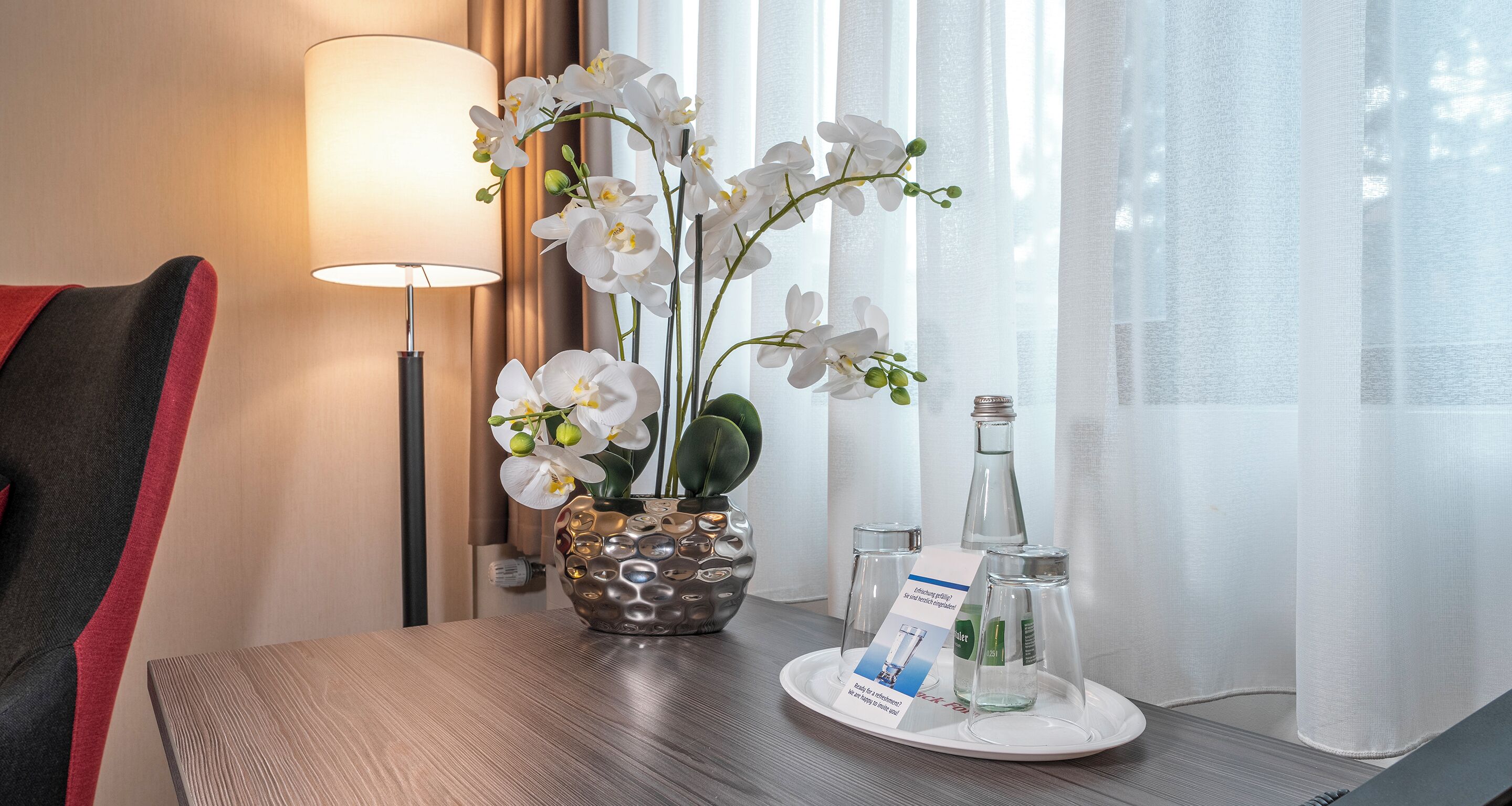 Habitación de hotel con una mesa de madera, una orquídea en maceta, una lámpara y una bandeja con una tarjeta y botellas de vidrio.