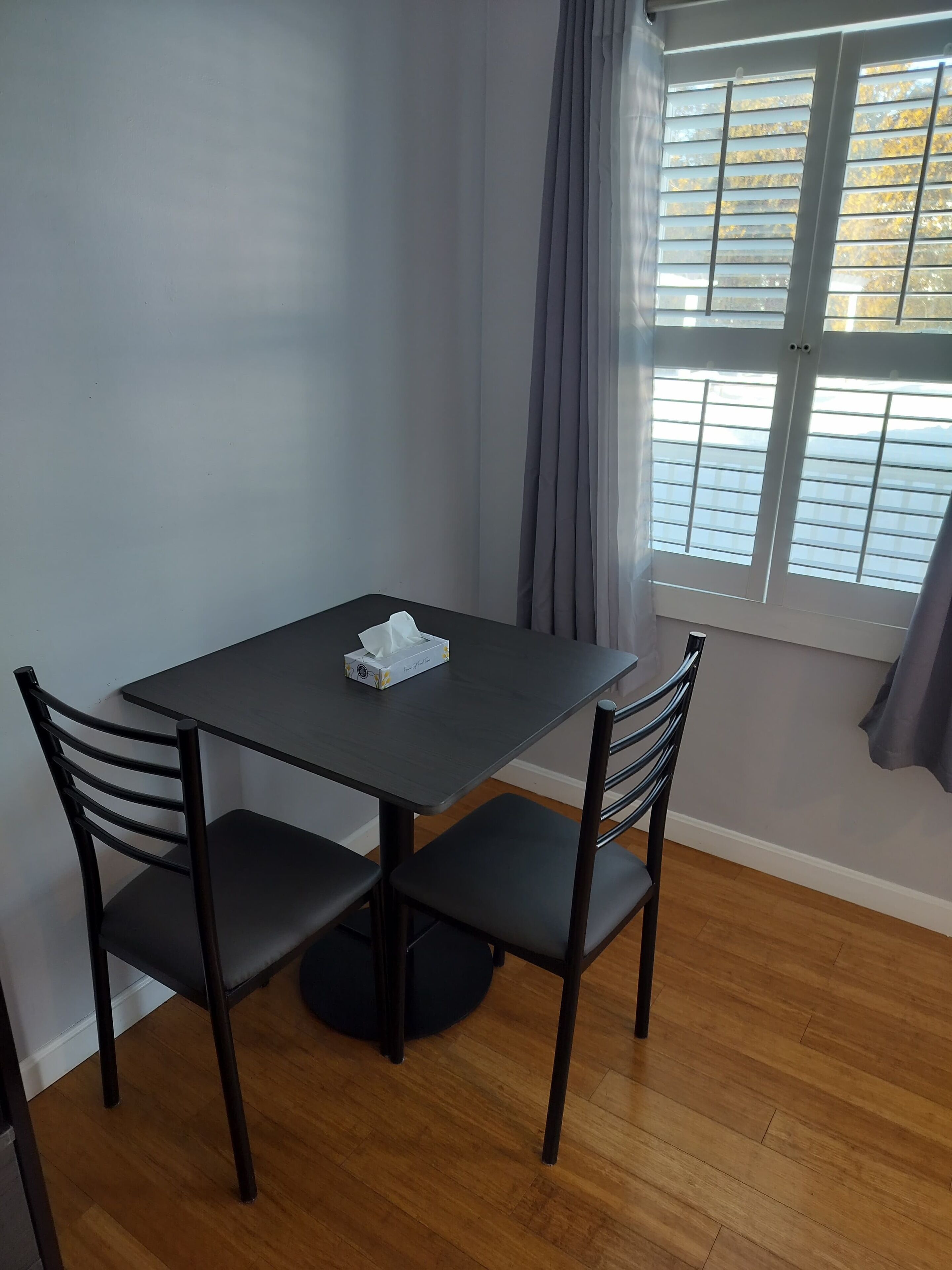 Un pequeño comedor con una mesa y dos sillas, una ventana con persianas y una cortina.