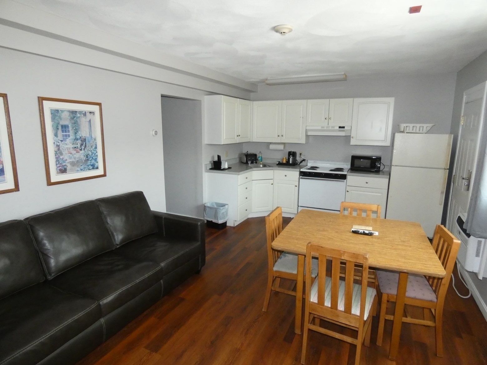 A compact living space with a kitchenette, a dining table with chairs, a black leather sofa, and framed pictures on the wall.
