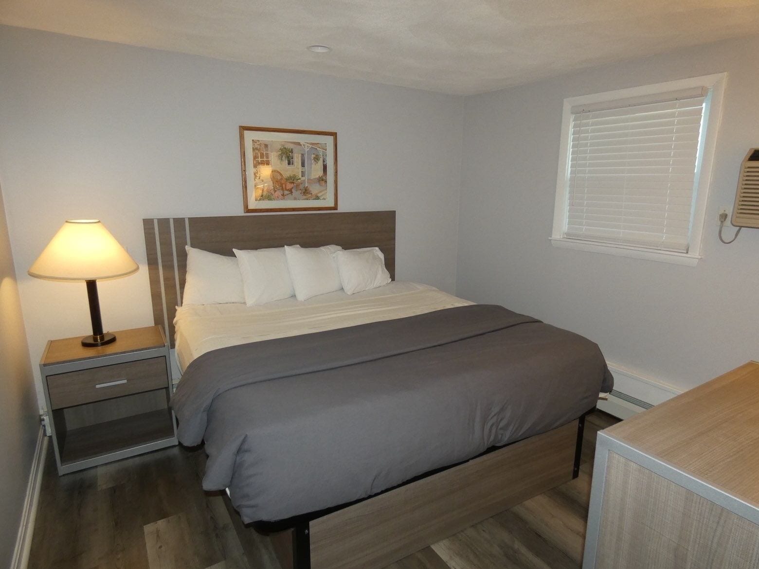 A bedroom with a bed, bedside table, lamp, and a window with blinds.