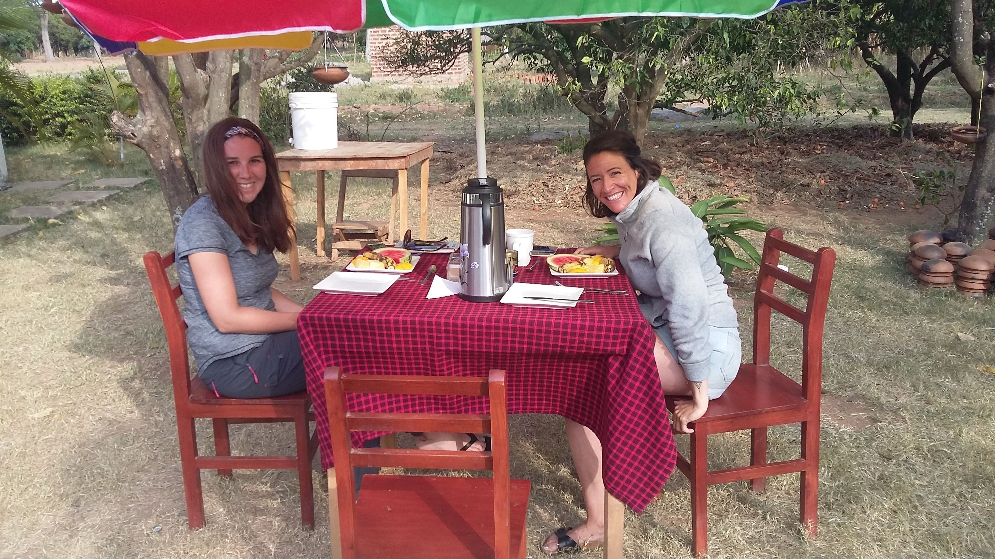 Two people seated at an outdoor table with a red checkered tablecloth, a metal coffee pot, and plates of food.