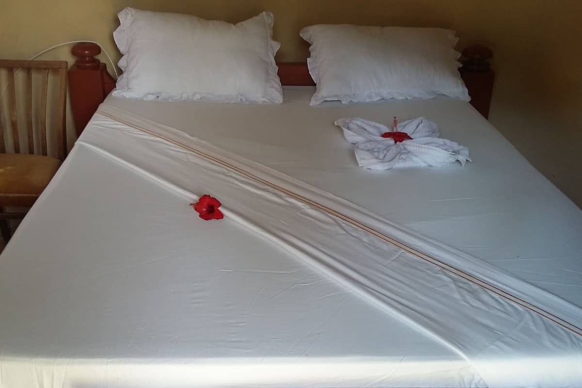 Un lit avec du linge de lit blanc, une chaise en bois et une fleur rouge posée sur le lit.