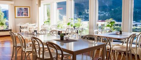 Un vaste espace dédié à la salle à manger, avec de nombreuses tables et chaises, de grandes fenêtres offrant une vue sur la ville et des plantes en pot.