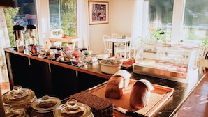 A breakfast buffet with various food items and condiments, a cutting board with bread, and a view of the outdoors through large windows. - F ørde Pensjonat (Sunnfjord)