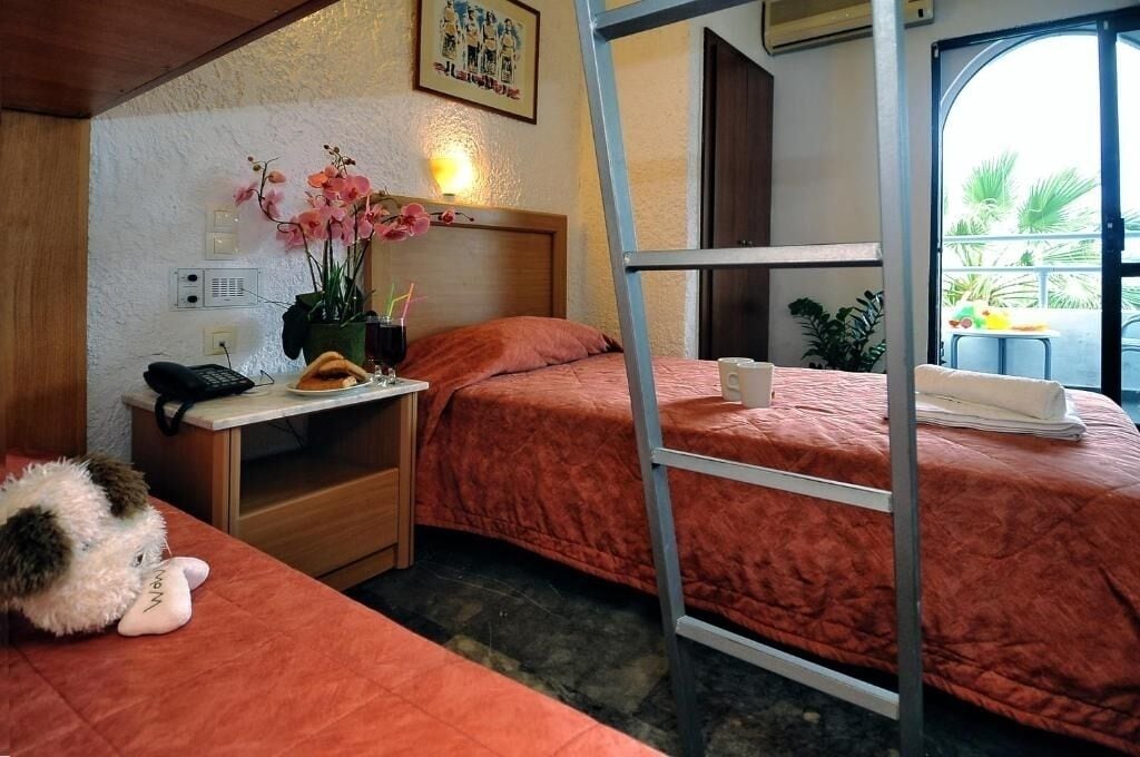 Una habitación de hotel compacta con dos camas, un escritorio con teléfono, una pequeña mesa con una planta y una ventana con vista a palmeras.