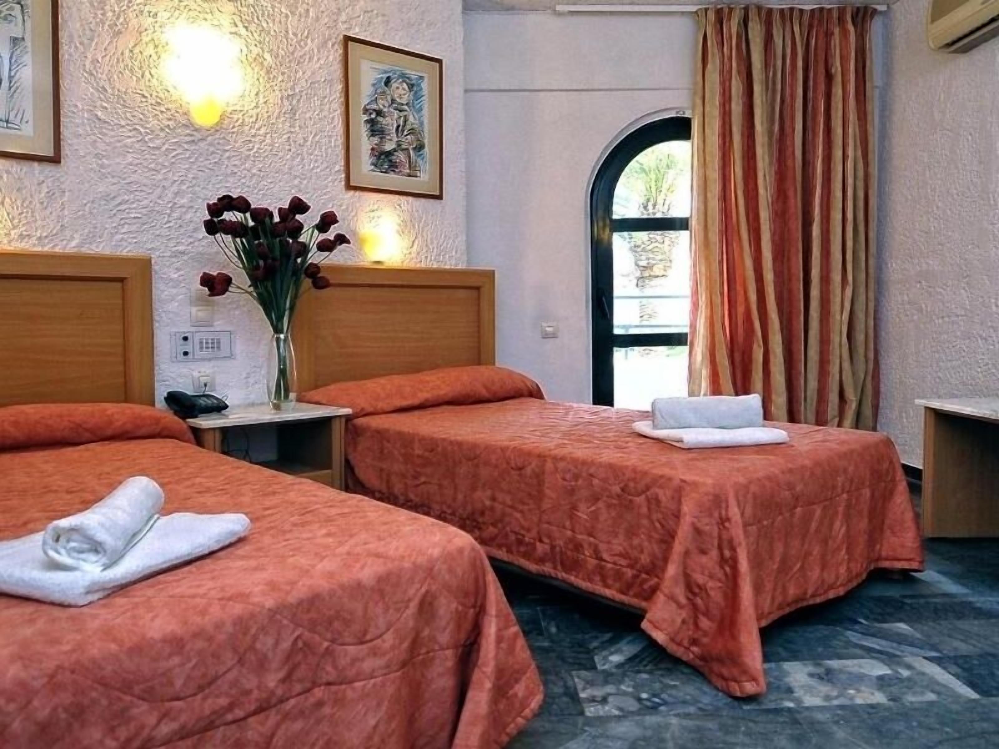 Habitación de hotel con dos camas, un jarrón con flores, un cuadro y vistas a través de una ventana.