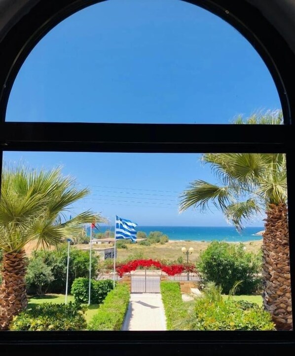 Vista a través de una ventana con cielo azul despejado, una bandera y un camino que lleva a una playa.