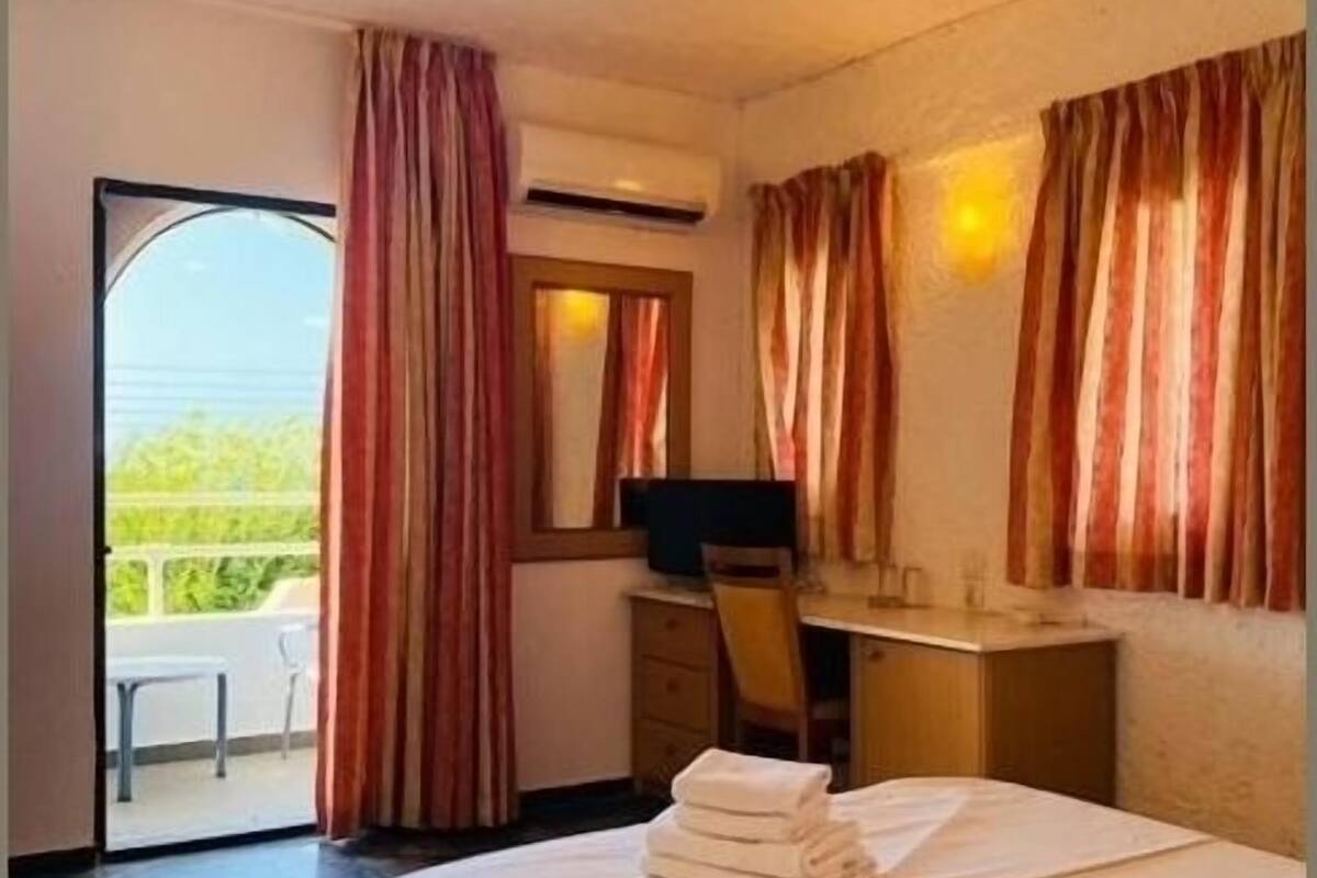 Habitació d'hotel amb un llit, una taula de treball amb un ordinador, una televisió i un balcó amb cortines.