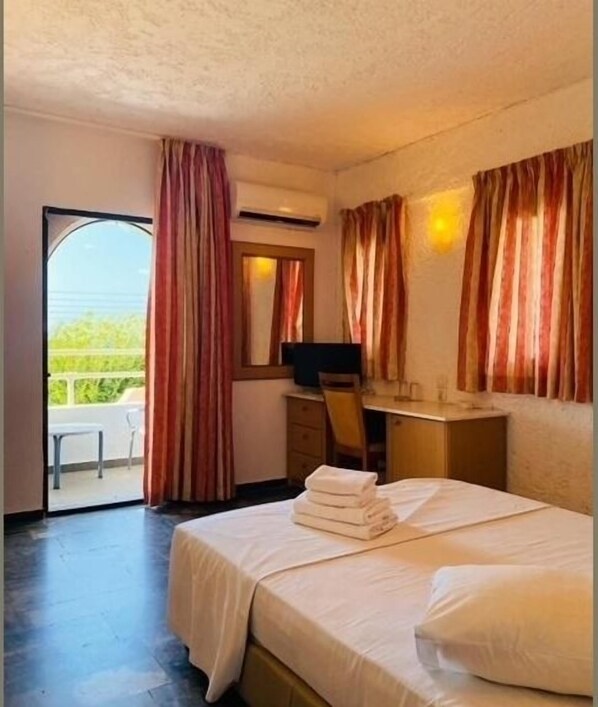 A hotel room with a bed, a desk with a computer, a TV, and a balcony with curtains. - Karteros Hotel (Hersonissos)