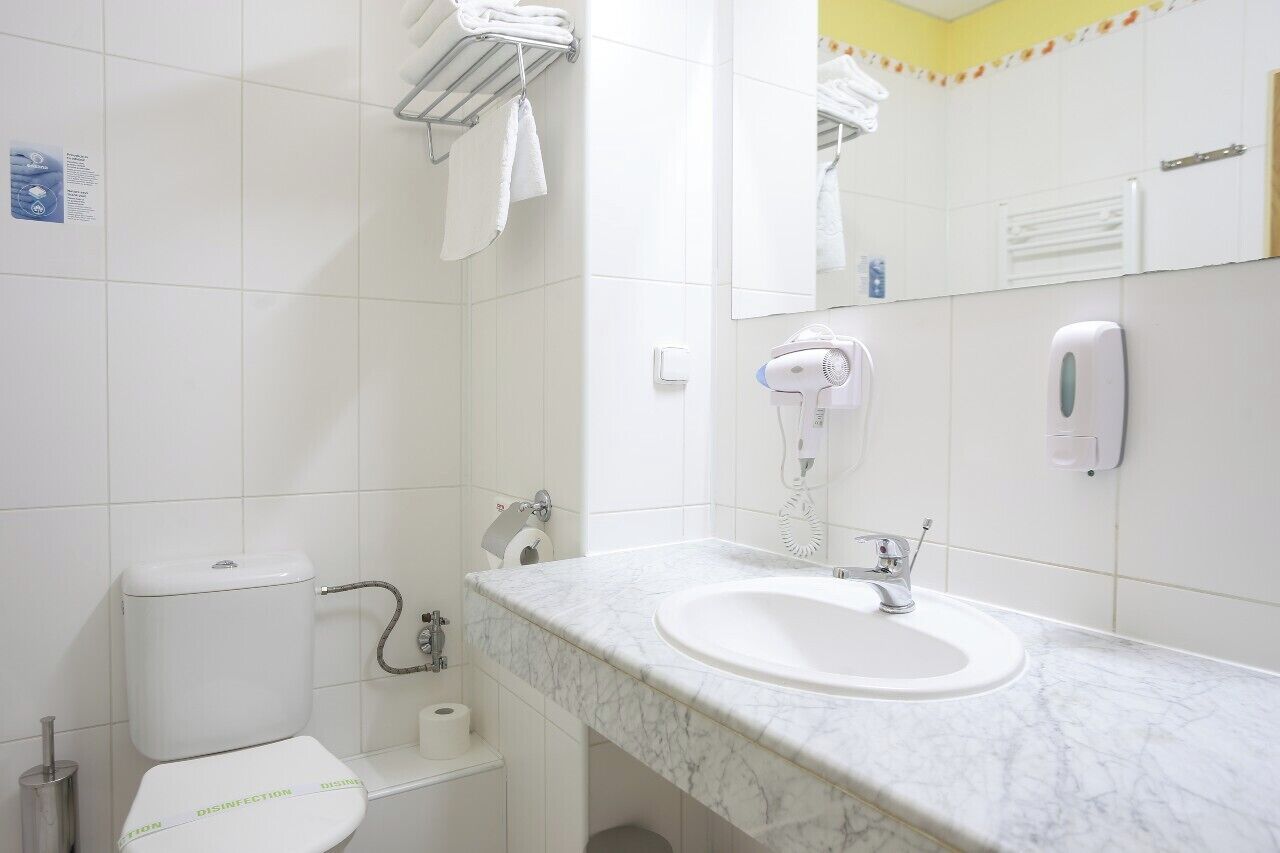 A bathroom with a white toilet, a sink with a marble countertop, a hairdryer, and a soap dispenser.