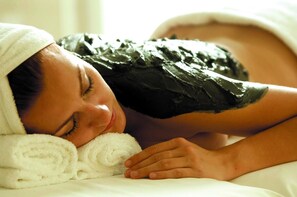 A person lying on a bed with a towel wrapped around their head, covered by a blanket.