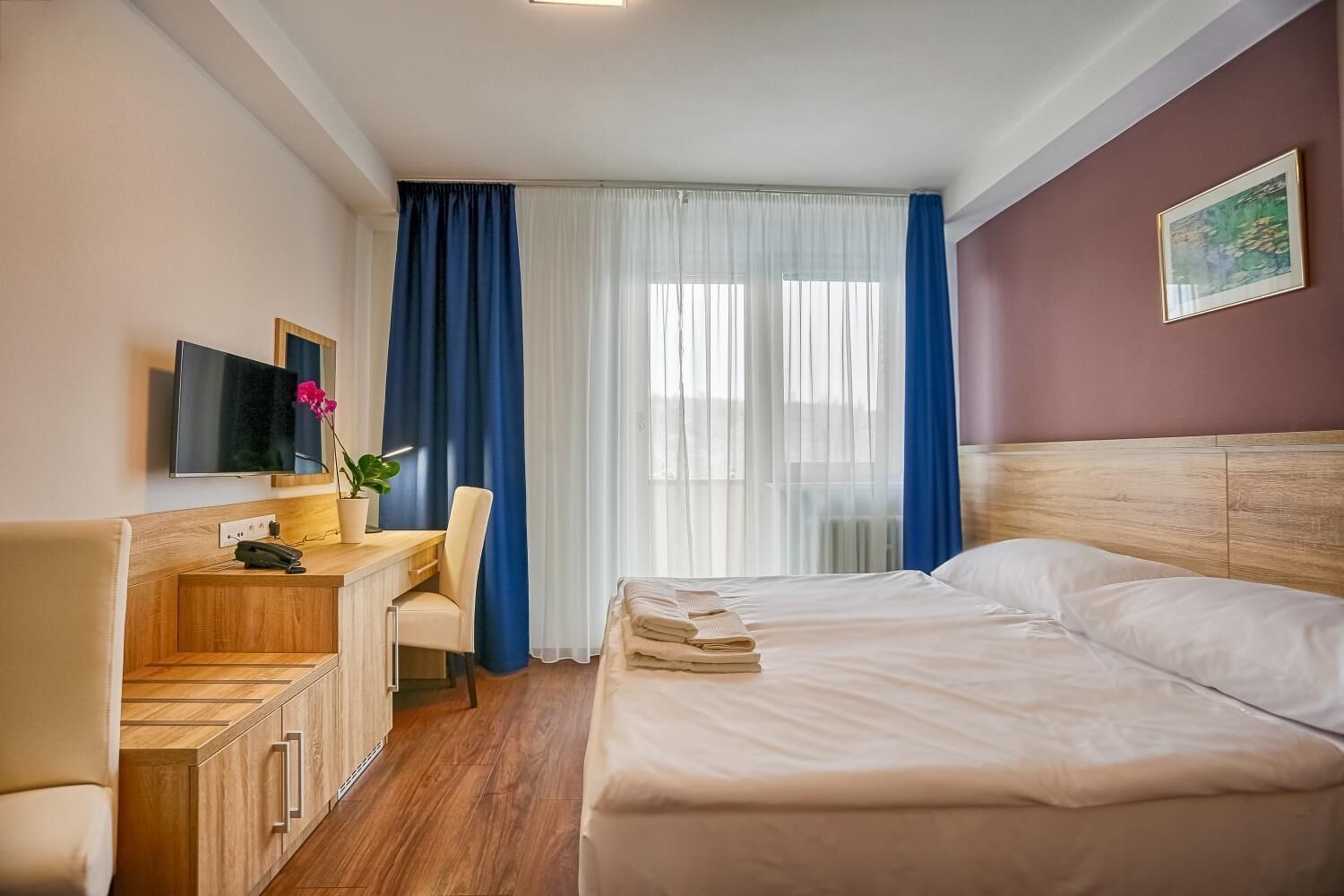 Una camera d'albergo con un letto, una scrivania con una sedia, una TV e una finestra con tende.