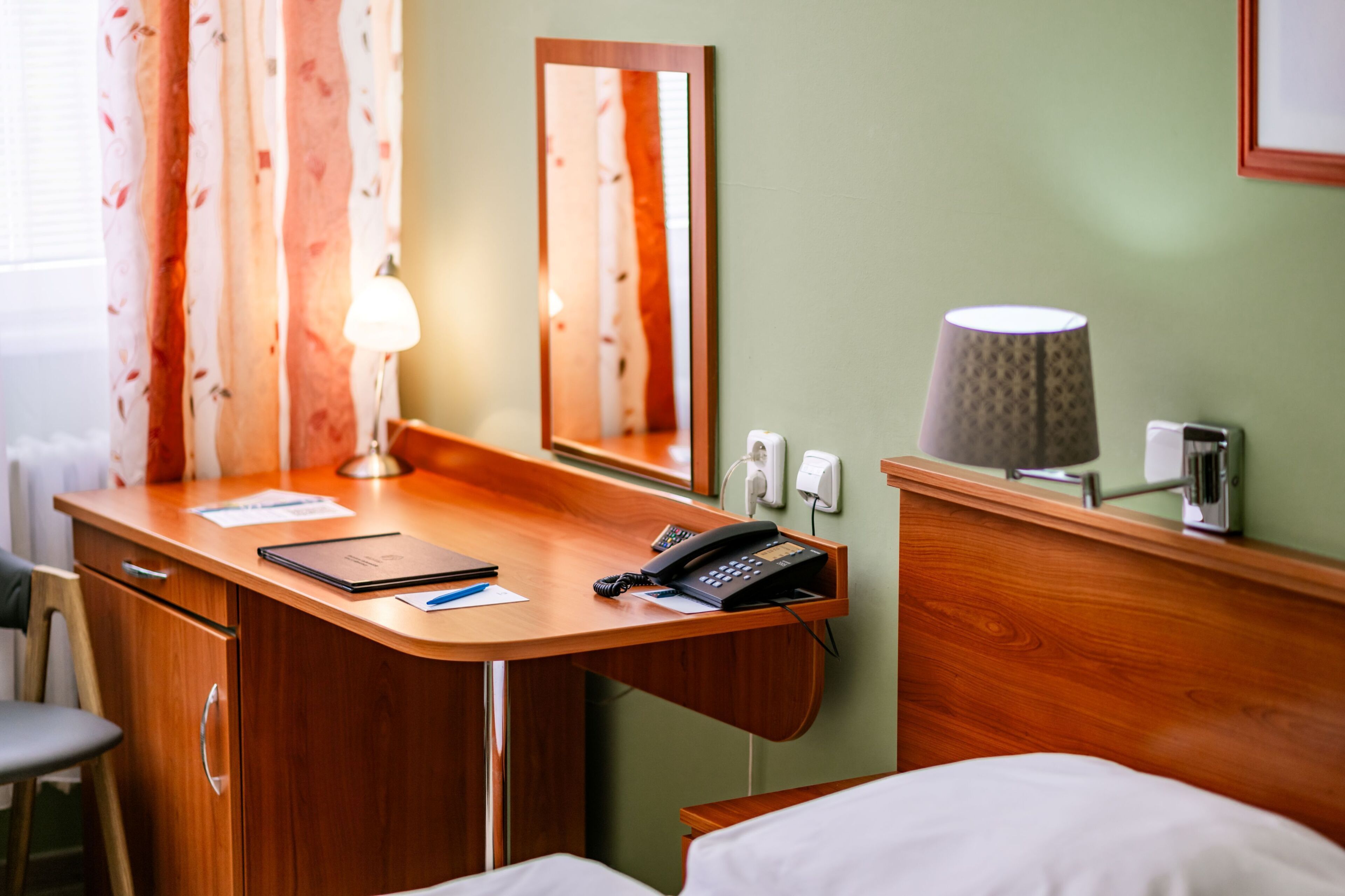A hotel room with a wooden desk, a telephone, a lamp, a mirror, and a bed.