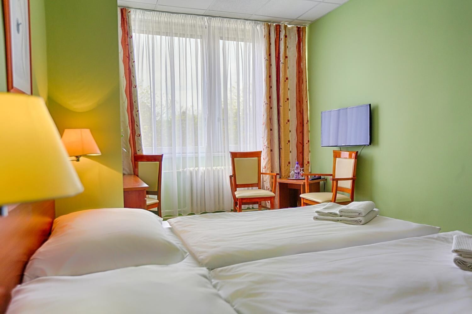 Une chambre d’hôtel avec deux lits, un bureau avec une chaise, un téléviseur fixé au mur et une fenêtre avec des rideaux.