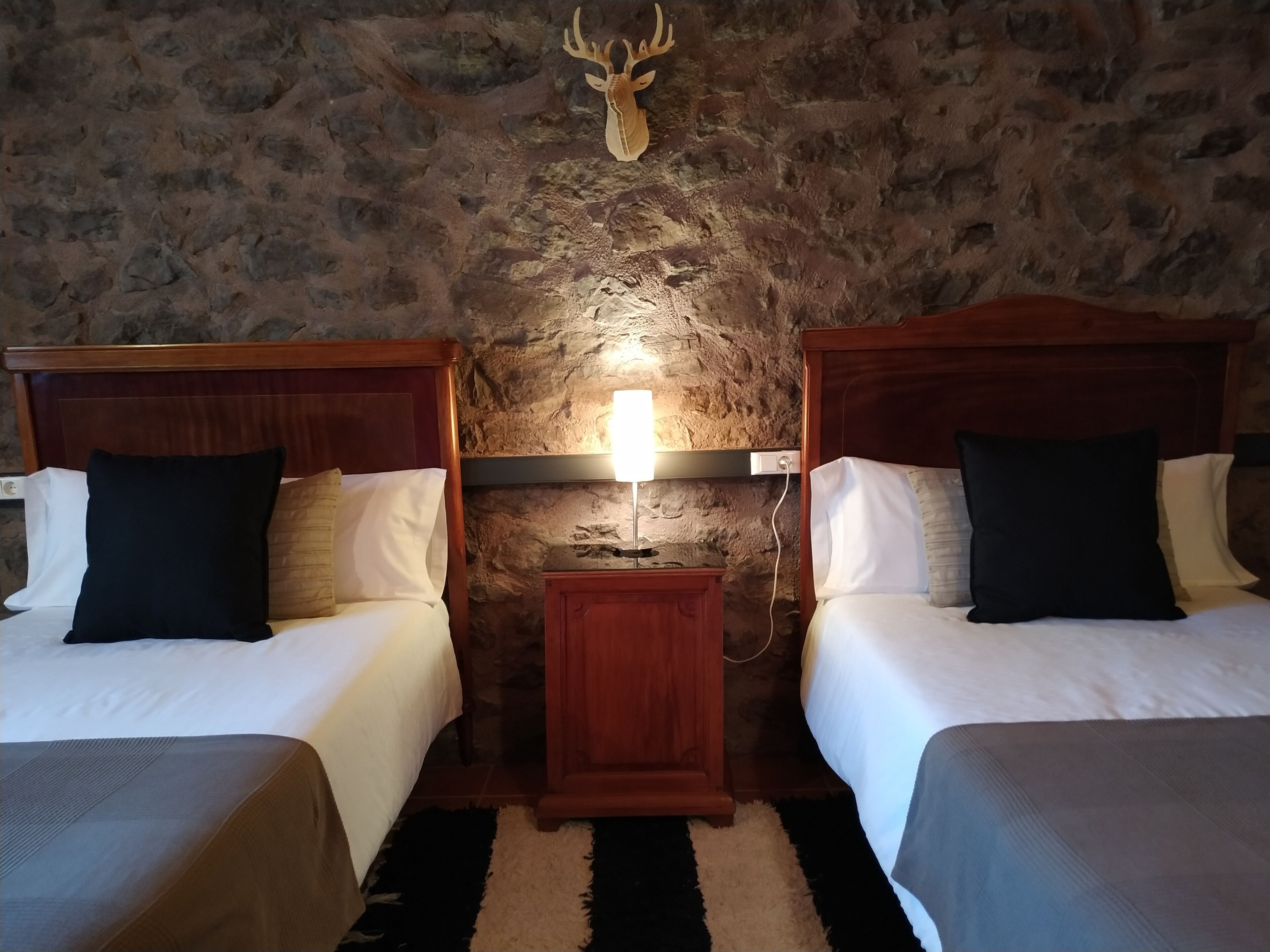 A hotel room with two beds, a stone wall, a mounted deer head, and a nightstand with a lamp.