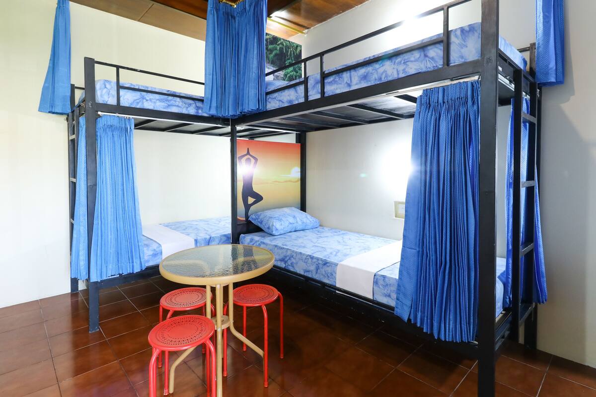 A bunk bed room with blue curtains, a round table with red stools, and a poster on the wall.
