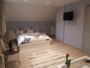A bedroom with a bed, a round rug, a small table with a lamp, a vase with a rose, and a TV mounted on the wall.