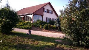 Une maison à deux étages, avec un toit recouvert de tuiles rouges, un porche abrité et un jardin bien entretenu, agrémenté de divers arbustes et d’un chemin menant à la maison.