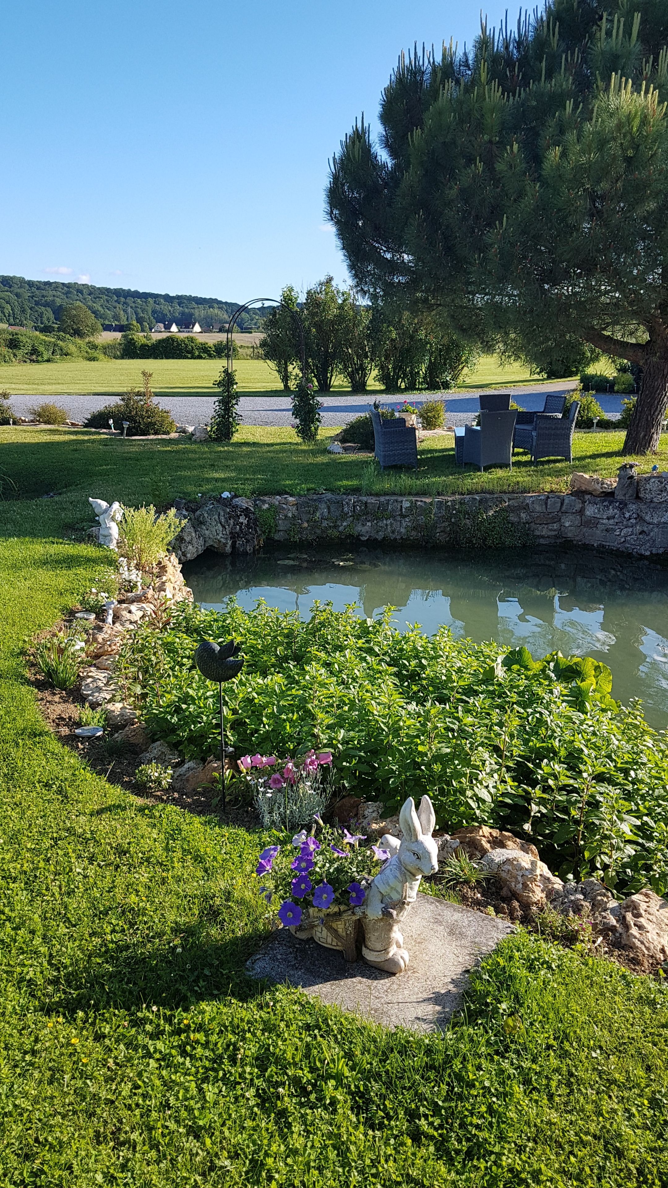 A serene garden with a pond, stone statues, and a seating area with chairs.