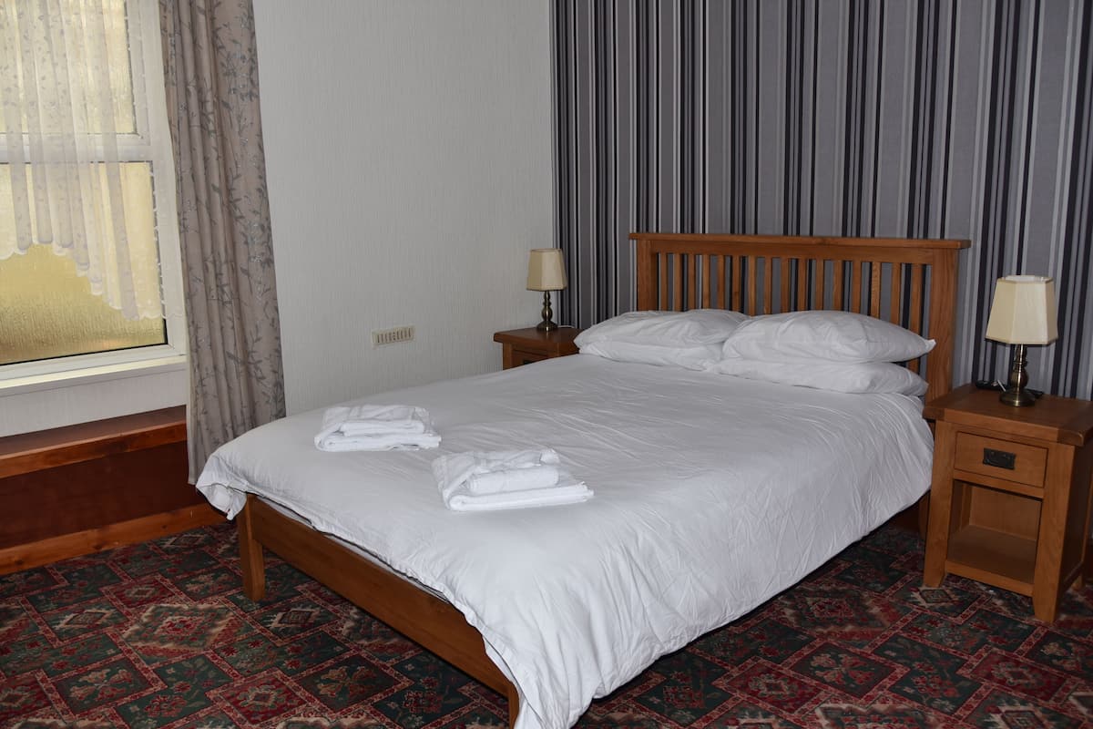 A neatly made bed with white linens and towels, a wooden headboard, two bedside tables with lamps, and a patterned carpet.
