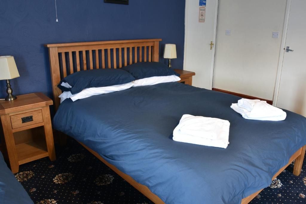 Een hotelkamer met een houten bed, twee nachtkastjes met lampen, een kledingkast en een donkerblauwe vloerbedekking.