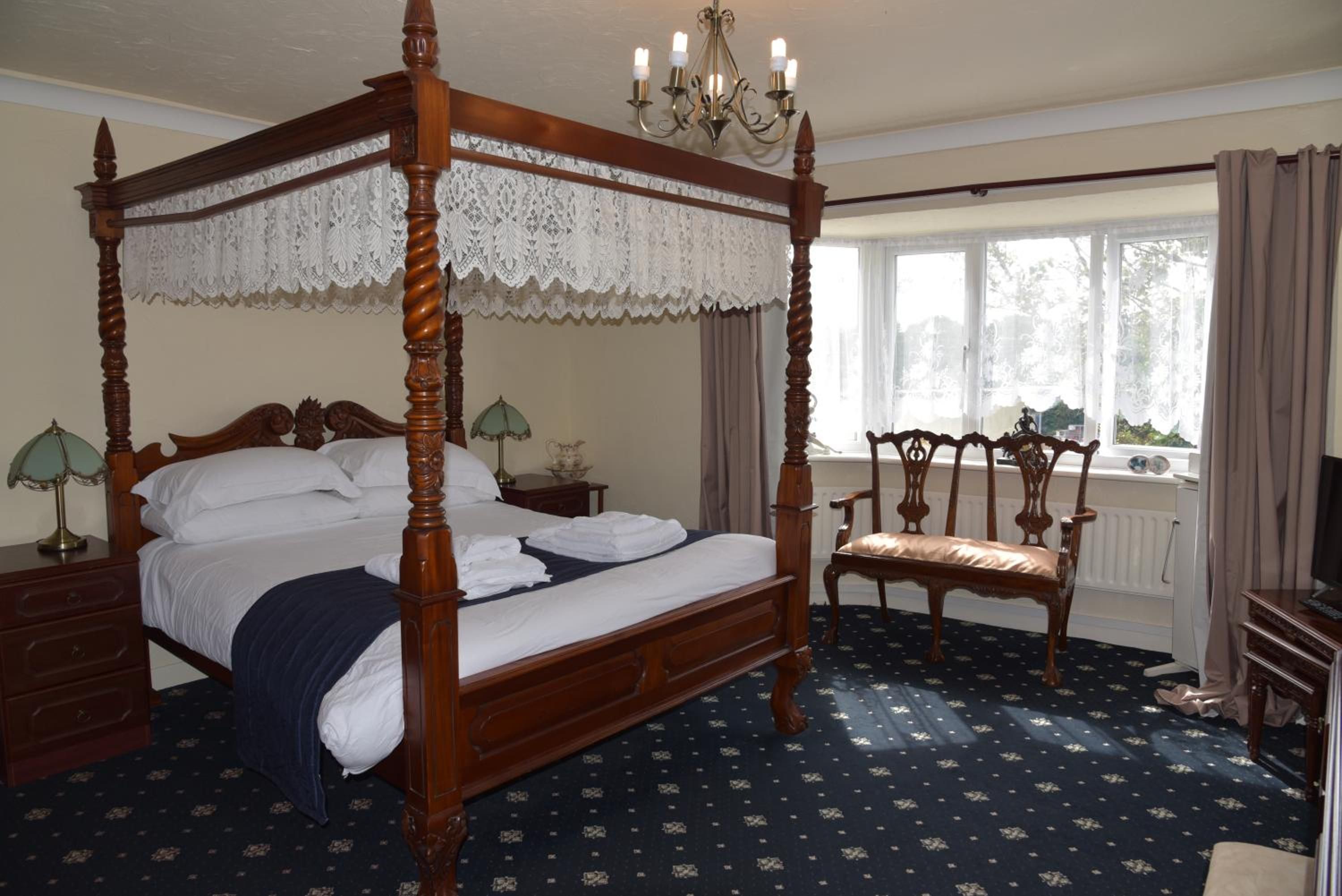 A four-poster bed with a lace canopy, a wooden chair, a nightstand with a lamp, and a window with curtains.