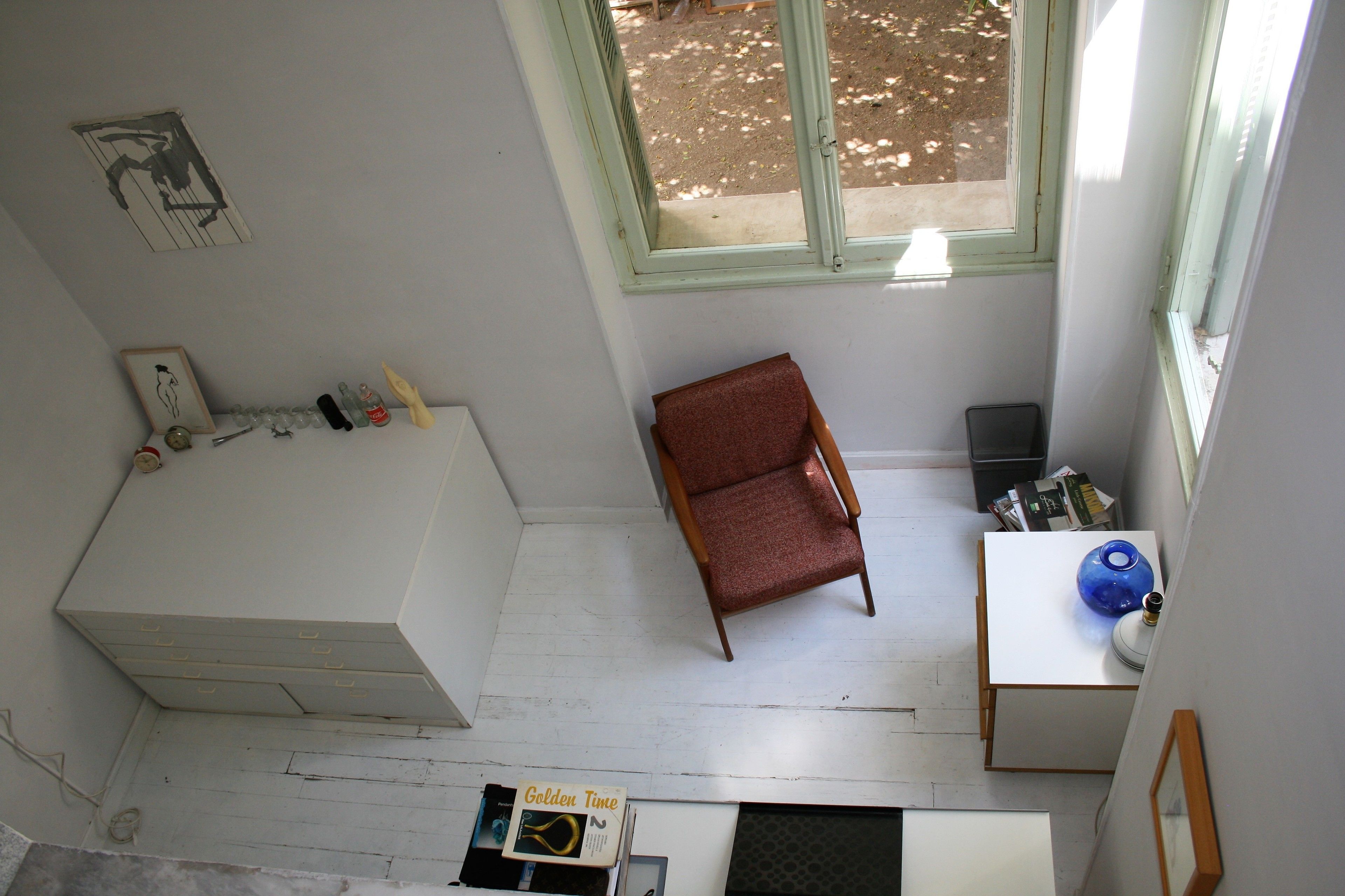 A room with a white bed, a red chair, a small table with a blue vase, and a window with a view of the outdoors.