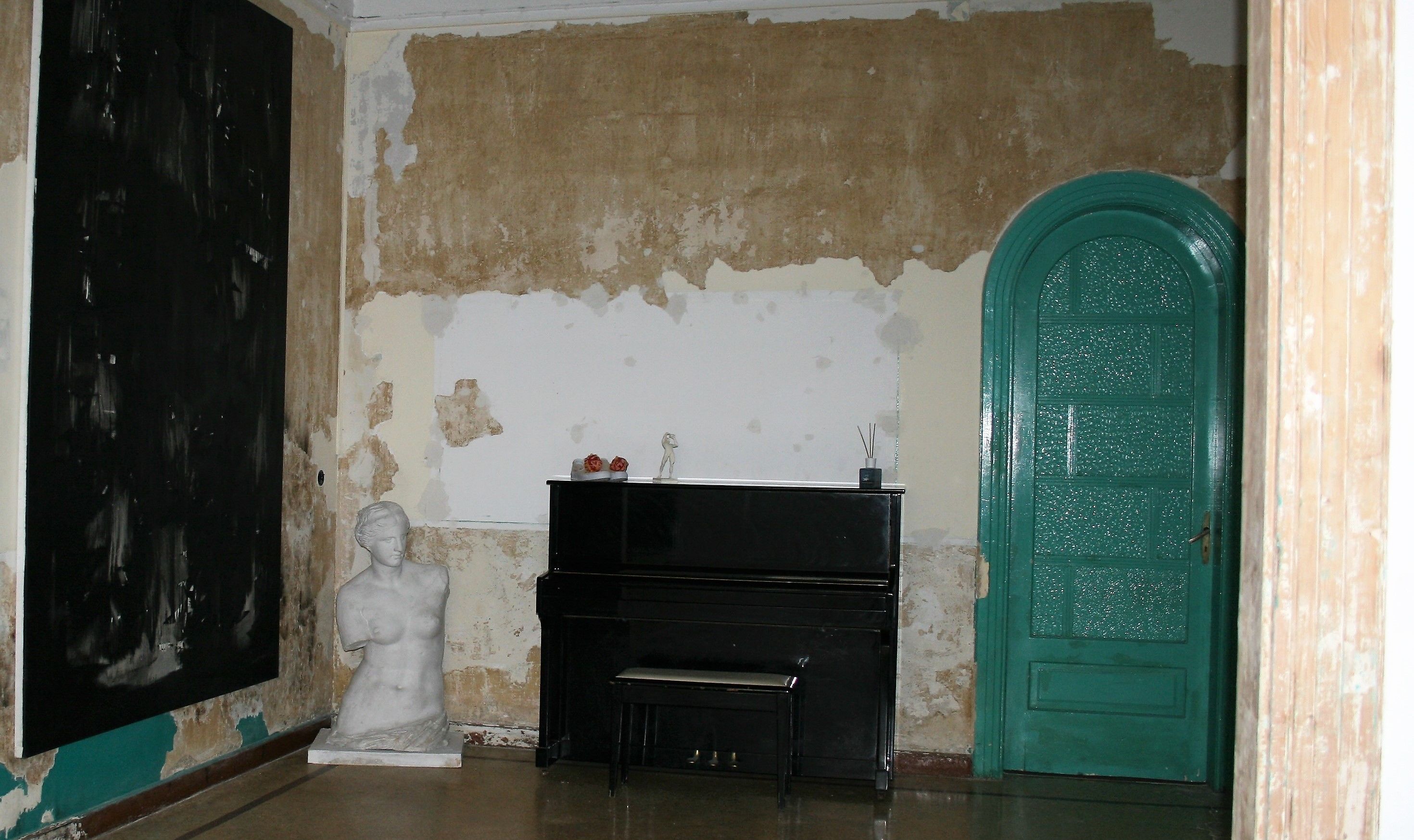 A room with peeling wall paint, a black piano, a white bust sculpture, and a green door.