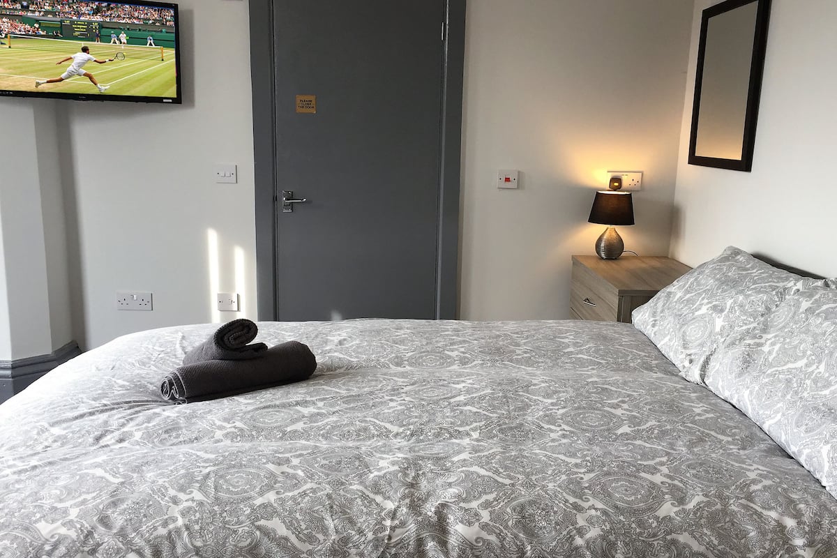 A hotel room with a bed, a television displaying a tennis match, a door, and a nightstand with a lamp.