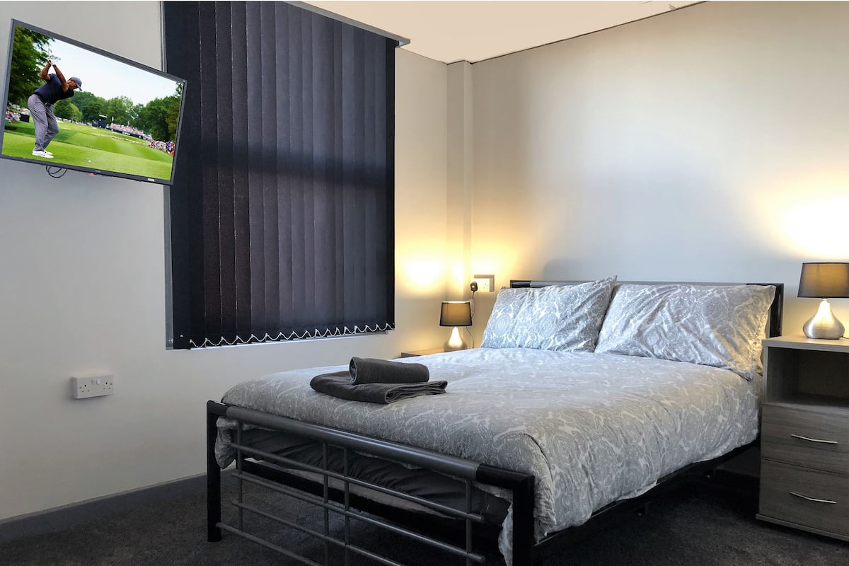 A bedroom with a bed, bedside tables, a TV mounted on the wall, and a window with blinds.