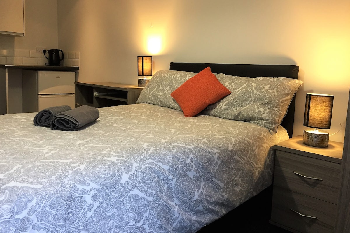 A neatly made bed with patterned bedding, a rolled towel, two bedside lamps, a small table with a coffee maker, and a nightstand with drawers.