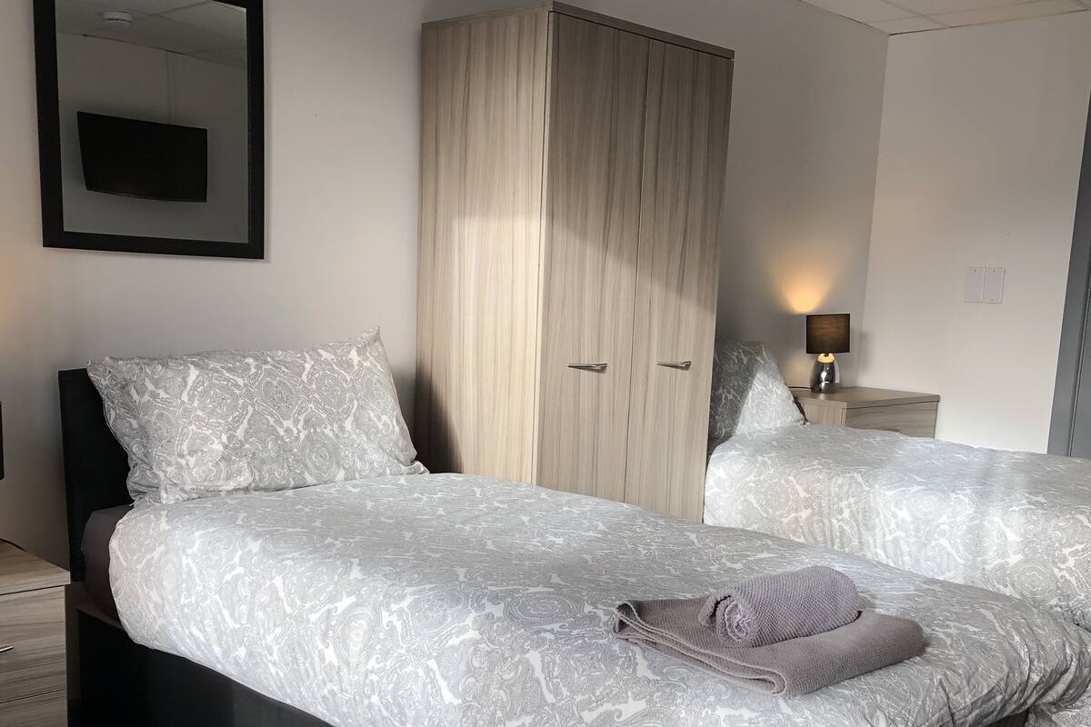A hotel room with two beds, a wardrobe, a bedside table with a lamp, and a mirror on the wall.