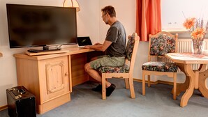 Un hombre trabaja en un escritorio de madera con una computadora portátil, un televisor y un altavoz. Hay una silla con motivos florales y una mesita con un florero.