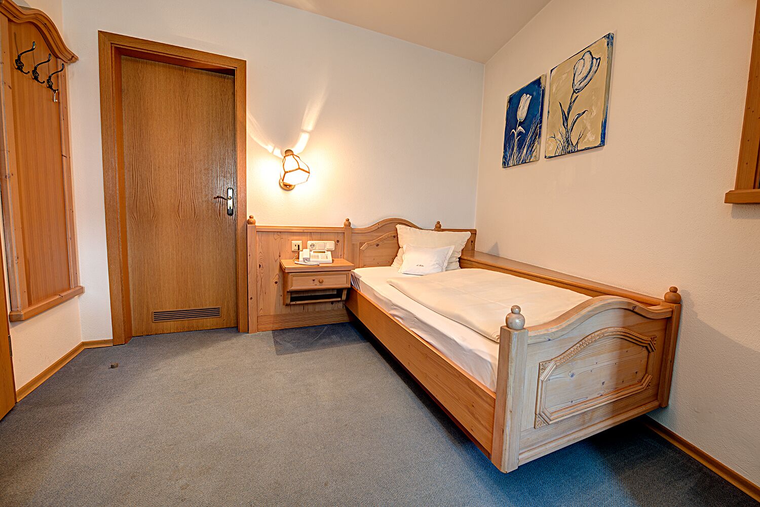 Un dormitorio con una cama de madera, una mesita de noche, una lámpara de pared y una puerta con perchero.
