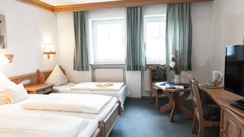 Habitación de hotel con dos camas individuales, un escritorio de madera con televisor, una silla, una mesa con un florero y una ventana con cortinas.