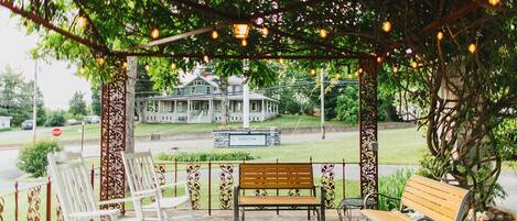 A covered outdoor seating area with stone flooring, white rocking chairs, and wooden benches.