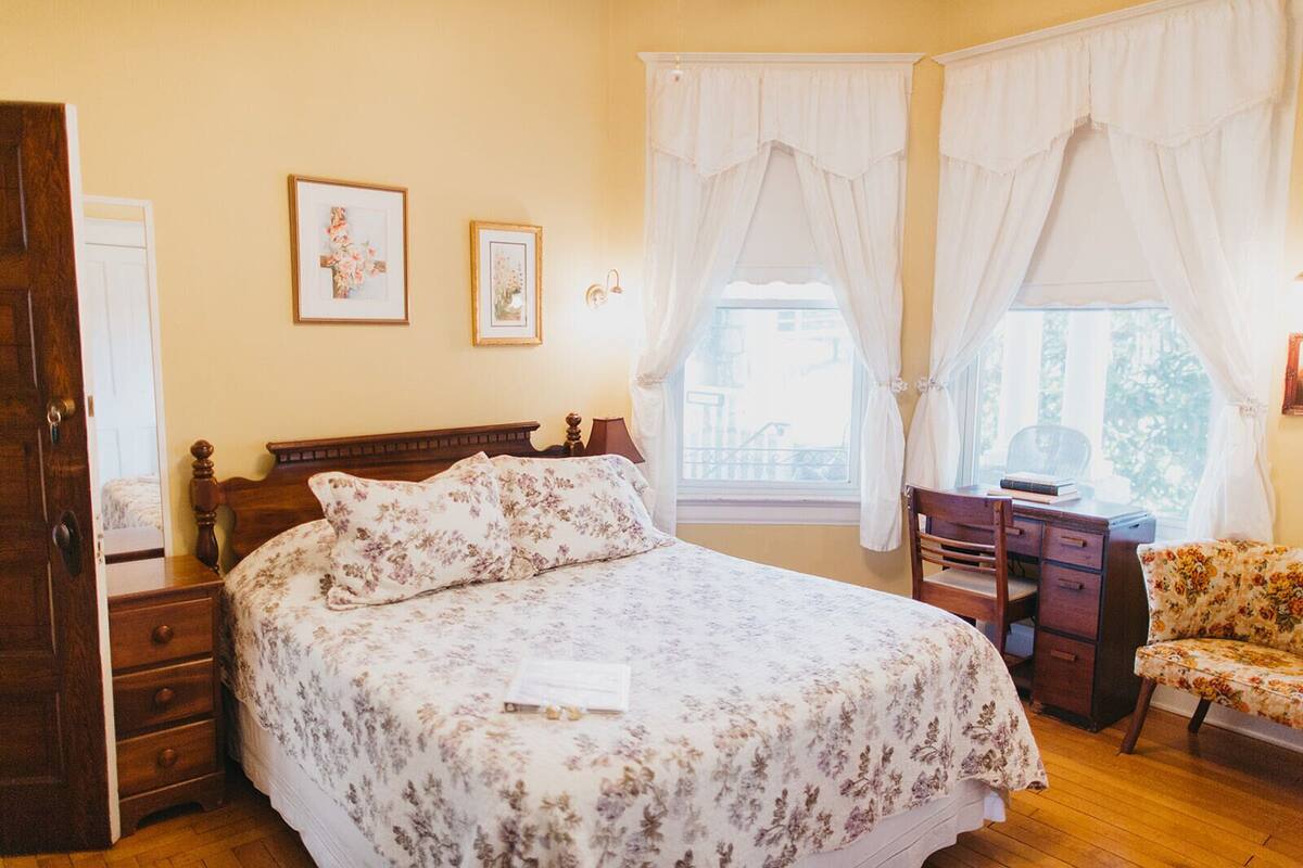 A bedroom with a wooden bed, floral bedding, a desk with a chair, and two framed pictures on the wall.