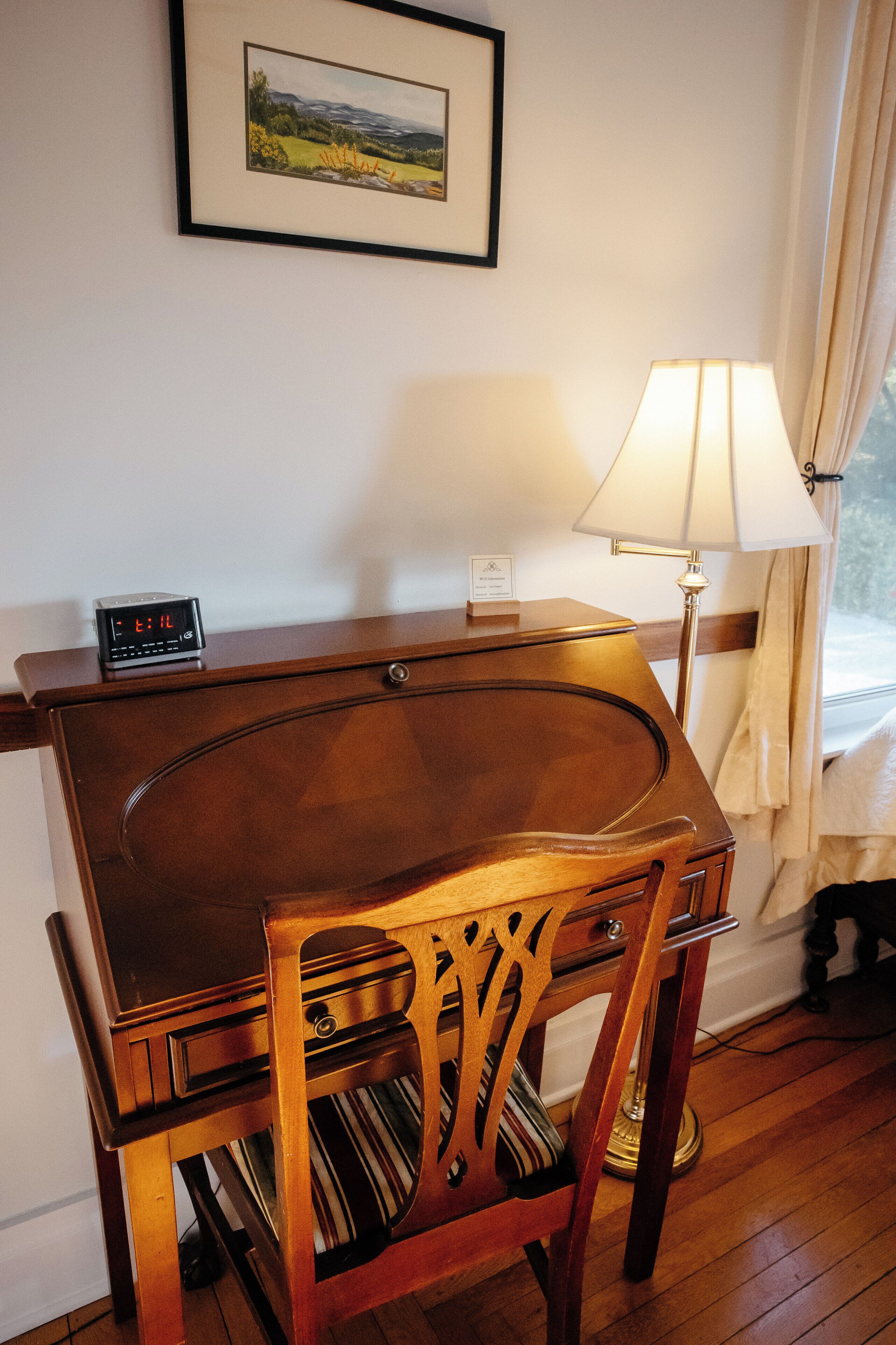 A wooden desk with a lamp, a clock, and a framed picture on the wall.