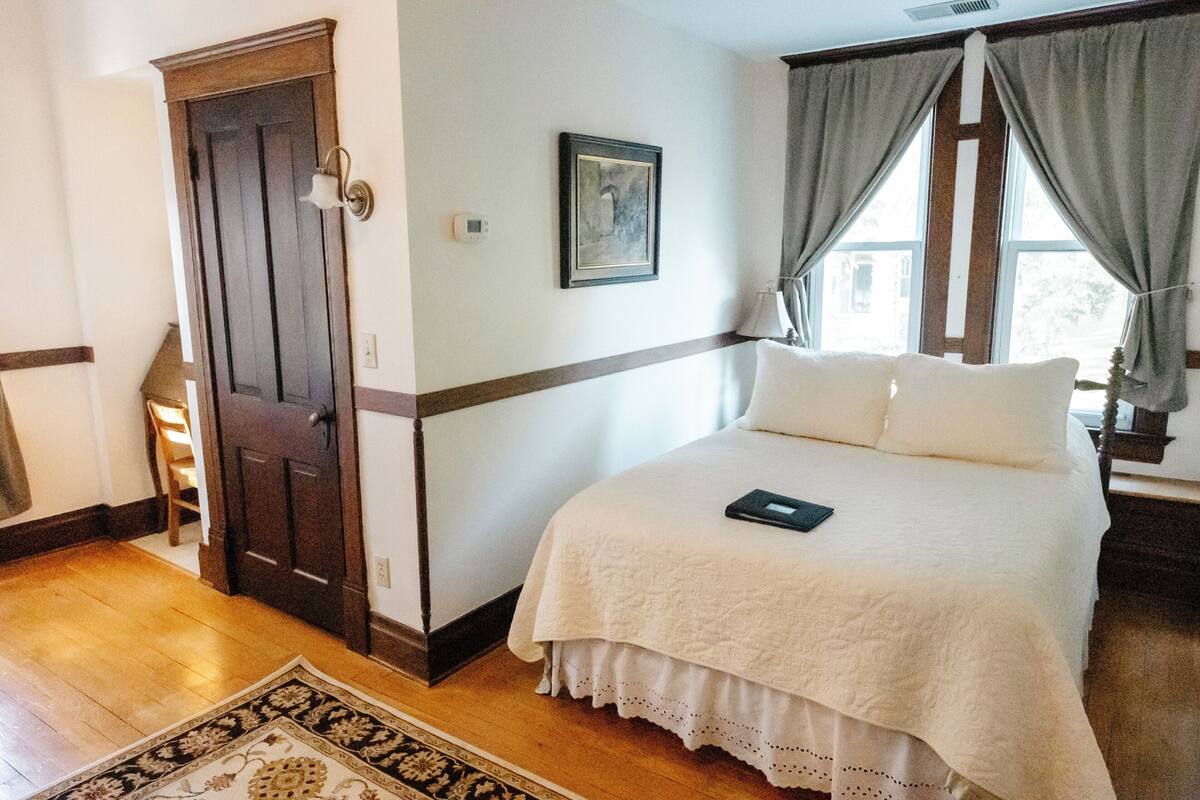 A bedroom with a wooden floor, a bed with white bedding, a door, a framed picture on the wall, and a window with curtains.