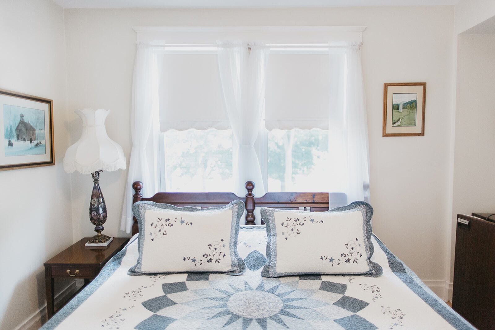 A bedroom with a bed, two pillows, a nightstand, a lamp, and a window with curtains.