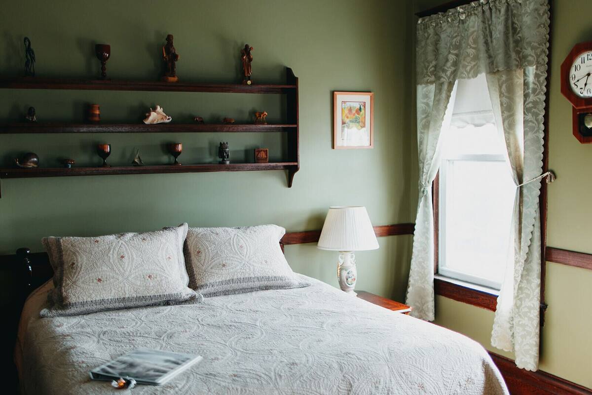 A bedroom with a bed, a nightstand, a lamp, a window with curtains, and shelves with decorative items.