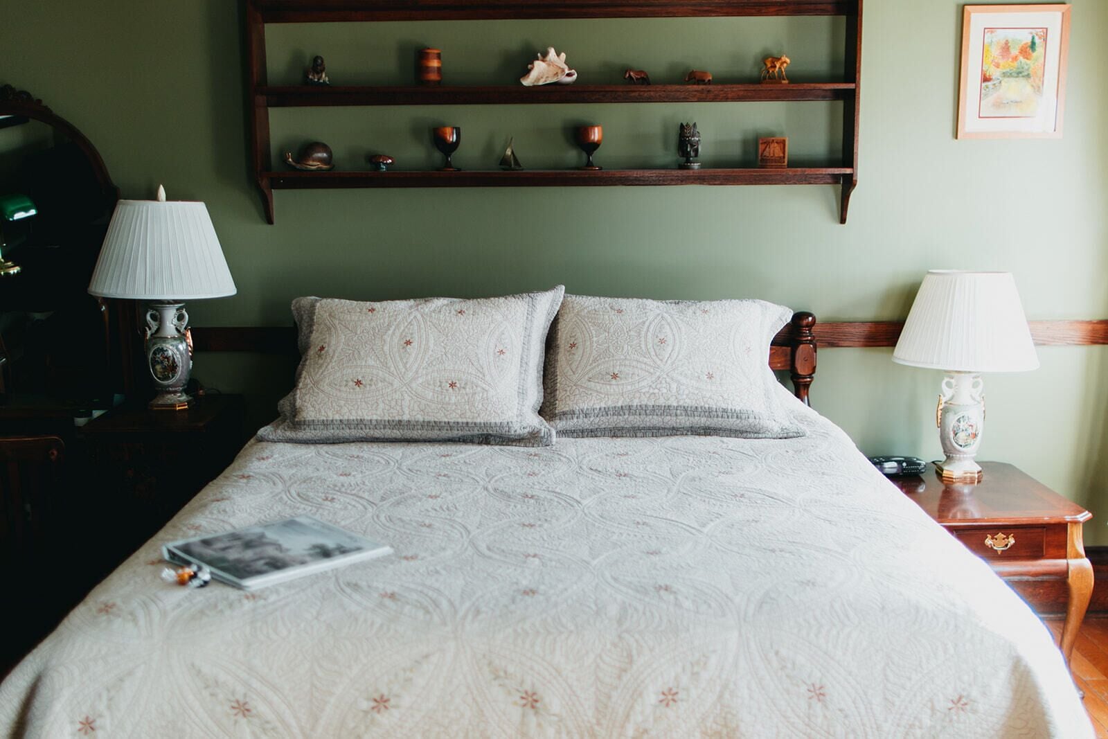 A neatly made bed with a patterned bedspread, two pillows, a bedside table with a lamp, and a shelf above the bed displaying various decorative items.