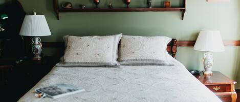 A neatly made bed with a patterned bedspread, two pillows, a bedside table with a lamp, and a shelf above the bed displaying various decorative items.