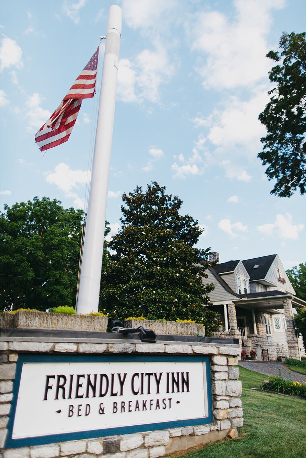A flagpole with an American flag, a stone sign for Friendly City Inn, and a house in the background.