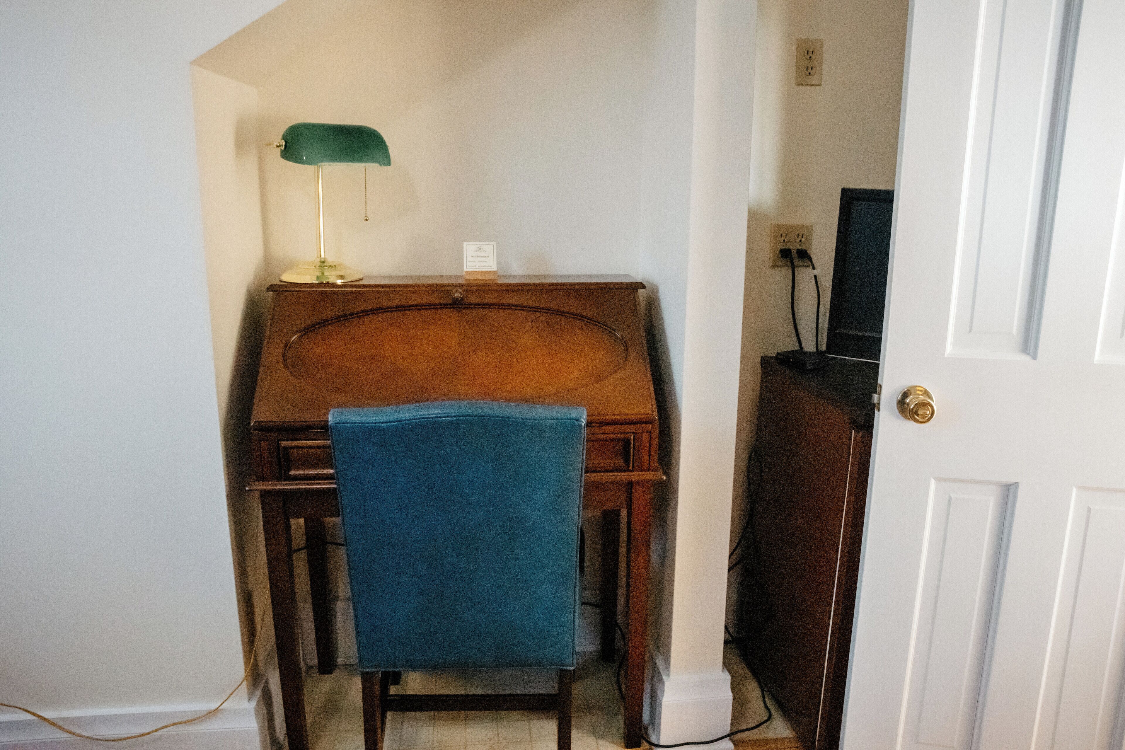 A room with a wooden desk, a blue chair, a green desk lamp, and a television on a cabinet.