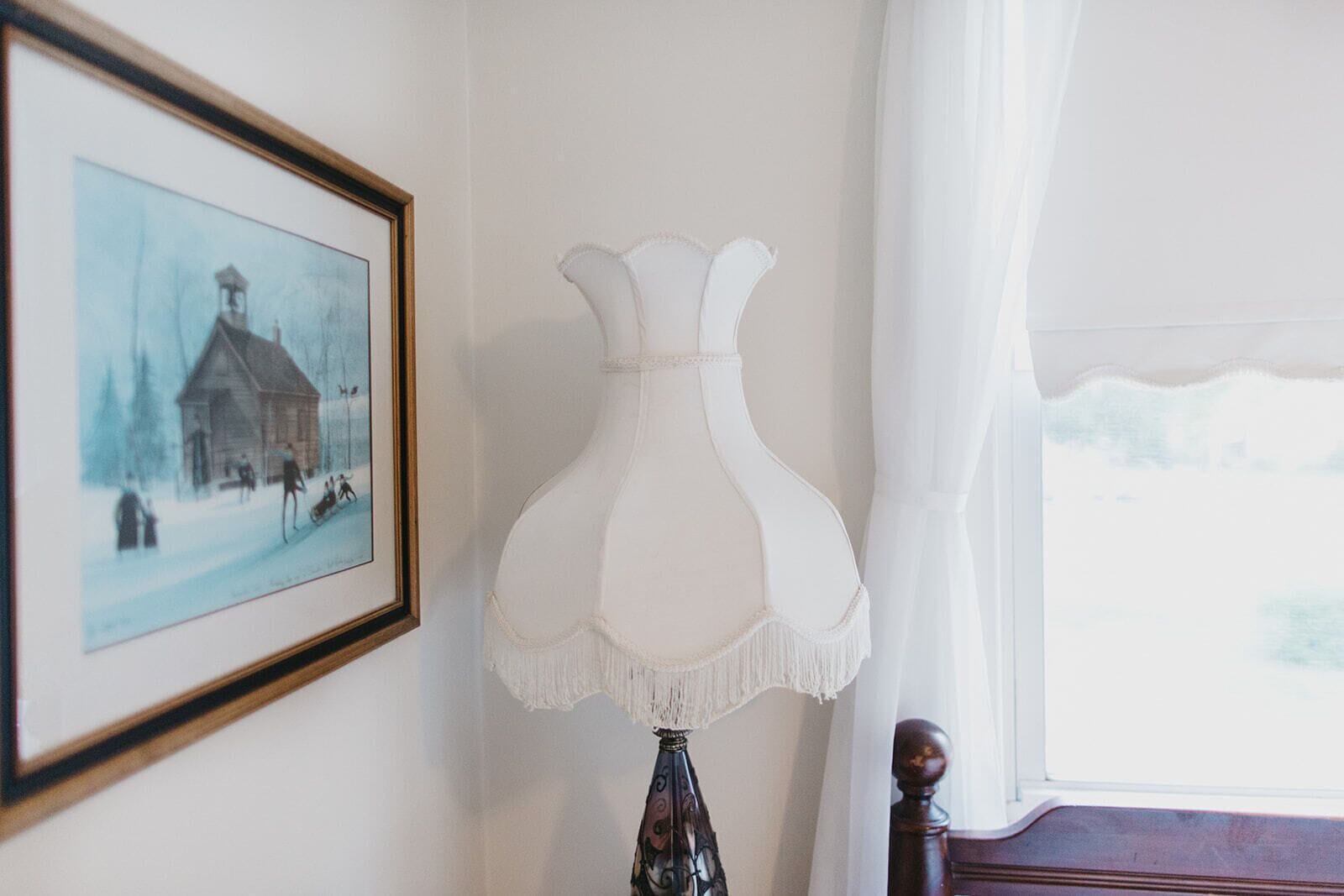 A room with a framed picture of a snowy scene, a white lamp, and a window with white curtains.