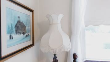 A room with a framed picture of a snowy scene, a white lamp, and a window with white curtains.
